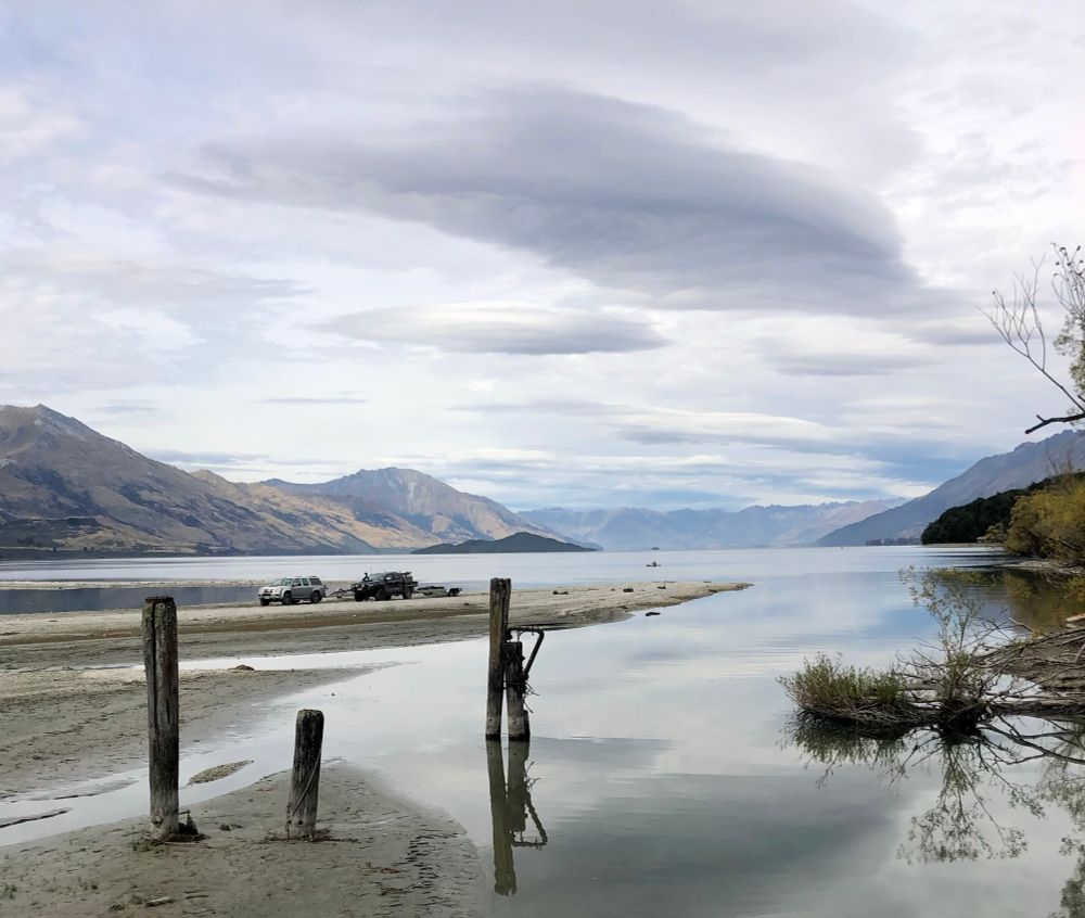 A calm dreamy blue and grey scene of a still reflective lake, with low sandbars and flax and wooden poles near the foreground, with low brown hills in the distance. 