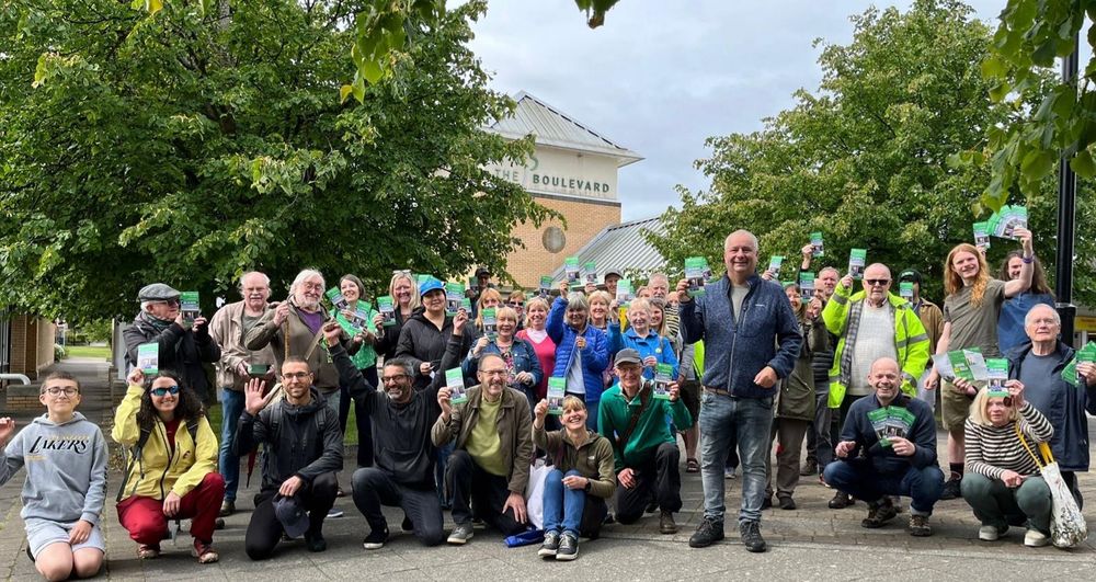 Crowd of 49 smiling activists posing with Green Party leaflets. Trees in the background.