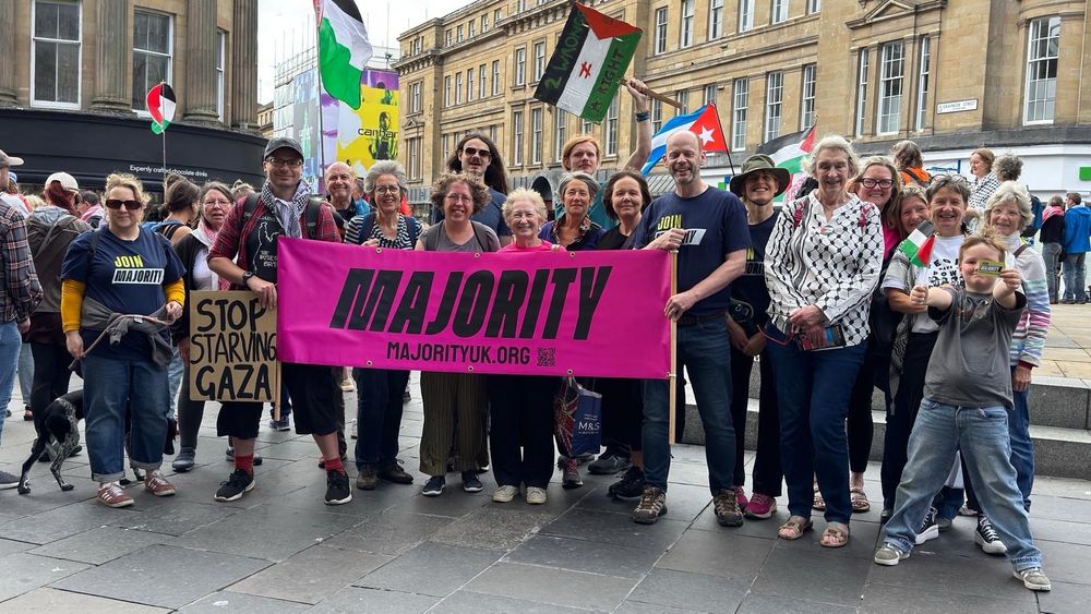 Picture of a group of 20-30 smiling people in Newcastle city centre with Palestine flags and placards. In front is a large pink banner with the words ‘Majority’ and ‘majority.org.uk’