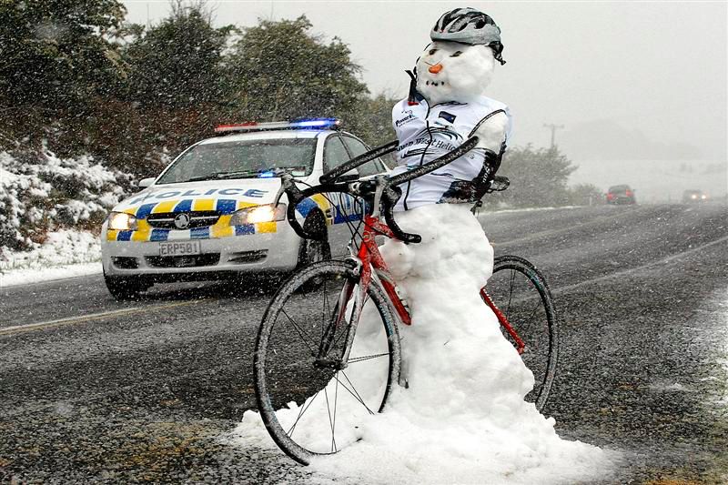 A snowman wearing a gilet and a helmet astride a red road bike. There's a police car in the background. It's snowing