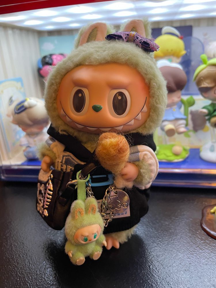 Another angle of Exciting macarons Labubu, Green Grape, in a cute jacket and purse with a tiny have a seat Labubu, QuQu, on her purse while she enjoys a needle felted fried chicken drumstick.