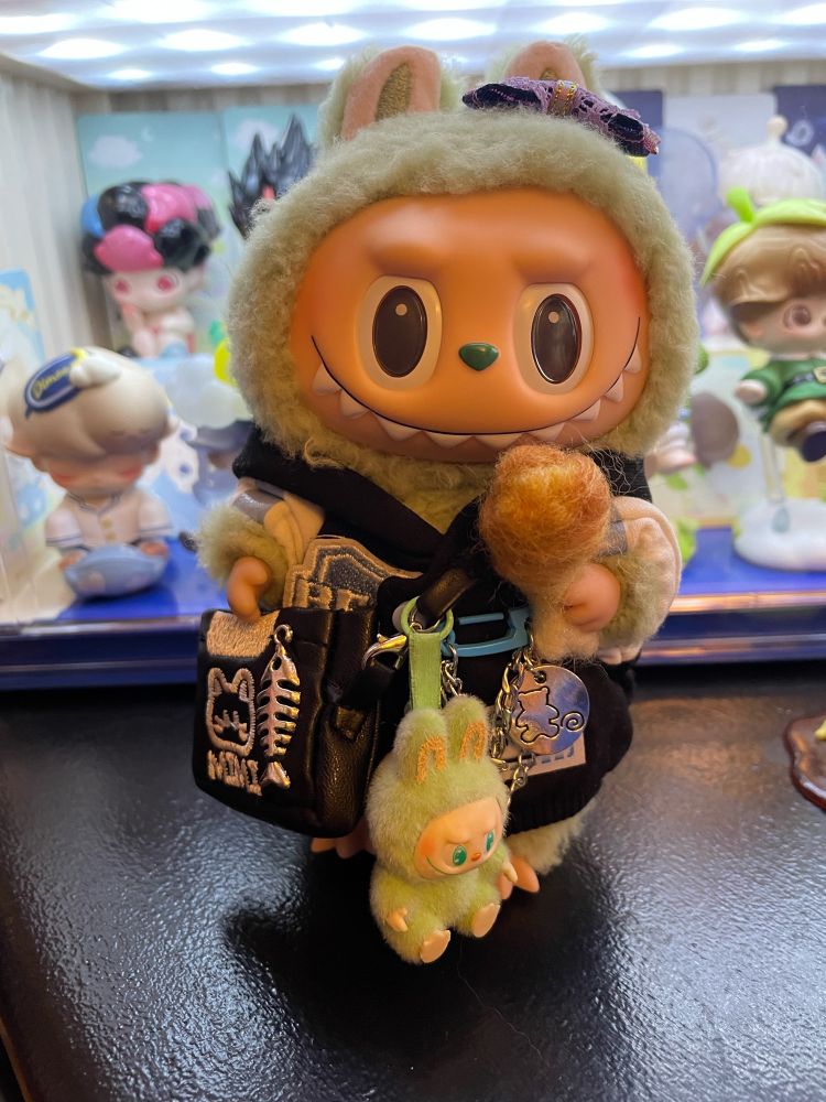 Exciting macarons Labubu, Green Grape, in a cute jacket and purse with a tiny have a seat Labubu, QuQu, on her purse while she enjoys a needle felted fried chicken drumstick.