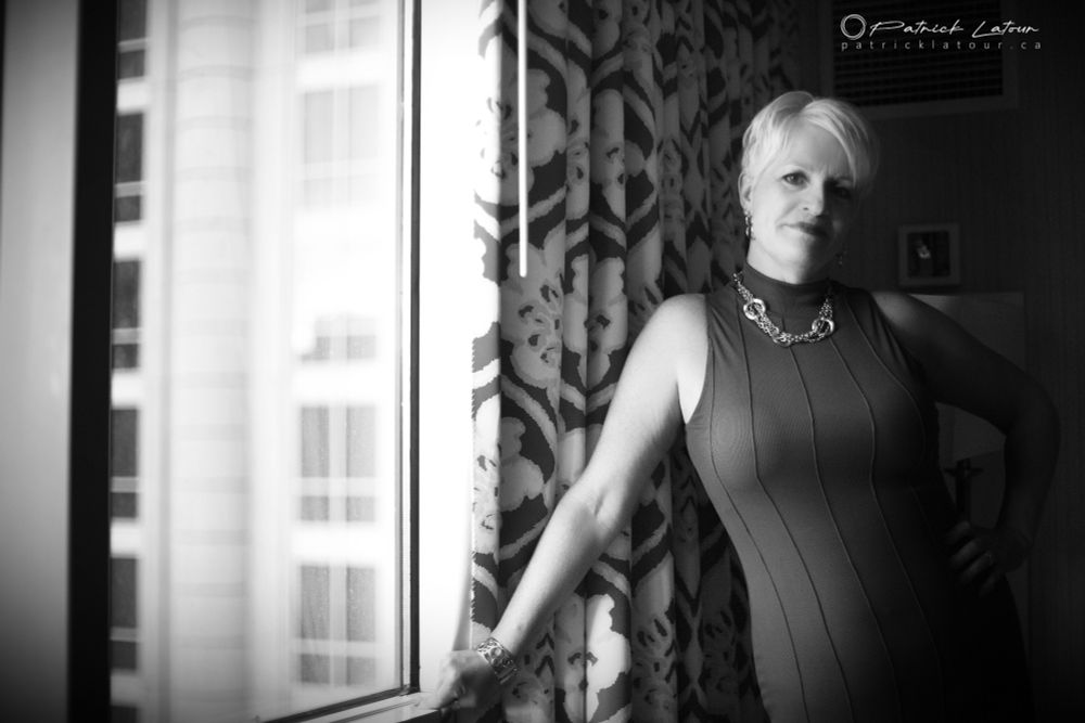 Elegant lady, with short blonde hair, in a dress by a window. The natural light arrive from the left and the right side is fading into the shadows.