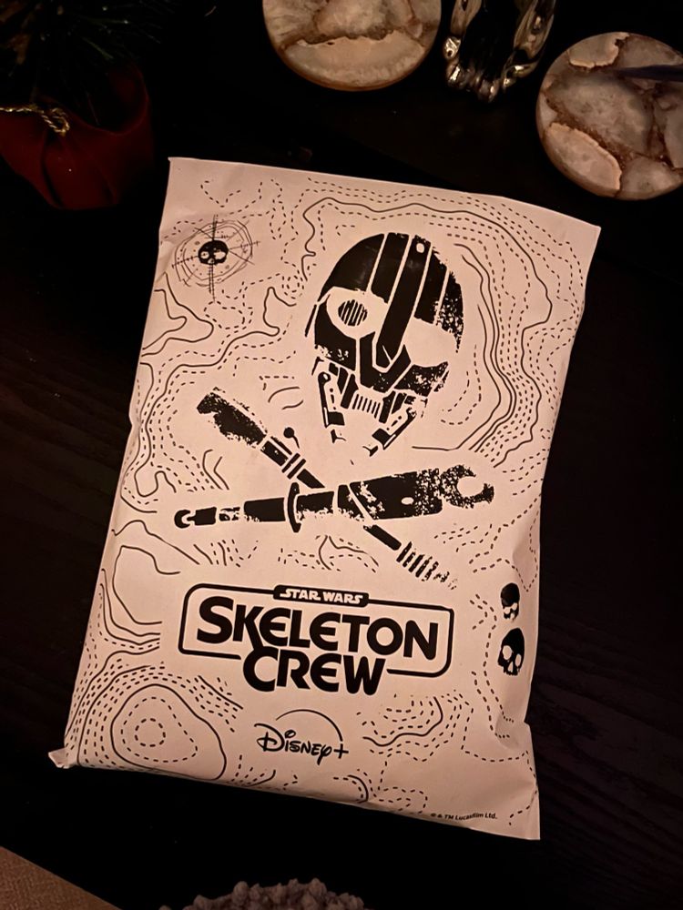 White paper bag with faint black dots over it like a map, there is a picture of SM-33s face and two objects crossed below it. It looks like a skull and cross bones. Below that are the words skeleton crew. 