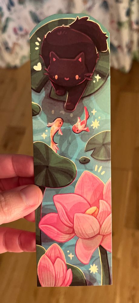 A glossy bookmark depicting a black cat standing on a lily pad in a pond. The cat’s paw is lightly touching the sparkly water and two koi circle around it between smaller lily pads. The cat has a white heart beside her head and can see the cat’s reflection in the water. In the foreground are two pink flowers. 
The illustration is by CC’s Art Cafe. 
