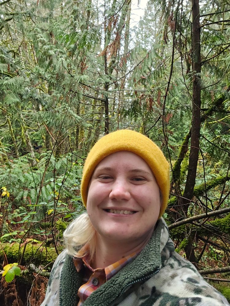 A selfie of me wearing a yellow beanie hat, orange flannel shirt, and wolf-print fleecy jacket. I'm standing in the middle of a lush Western hemlock swamp in my friend's parents' back yard.