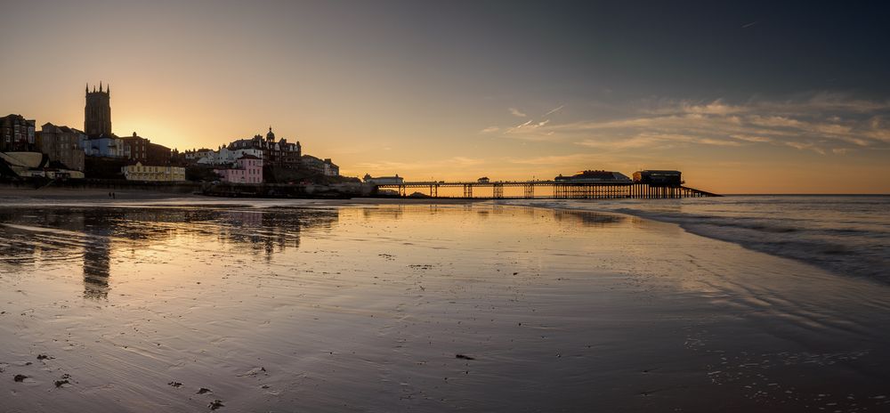 The town of Cromer and its Pier stretching into the sea reflected on a wet beach as the sun sets behind.