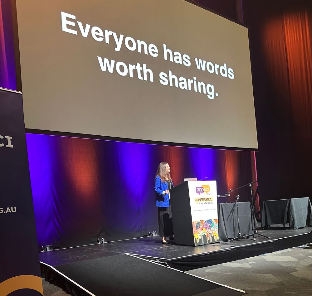Jordyn, standing at the podium as she delivers her keynote during the AGOSCI conference. On screen there’s a message in white lettering, “Everyone has words worth sharing.”