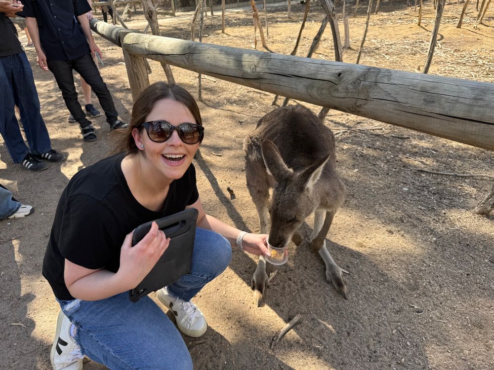 Jordyn, knelt down next to a kangaroo, holding a cup in her left hand to feed it.