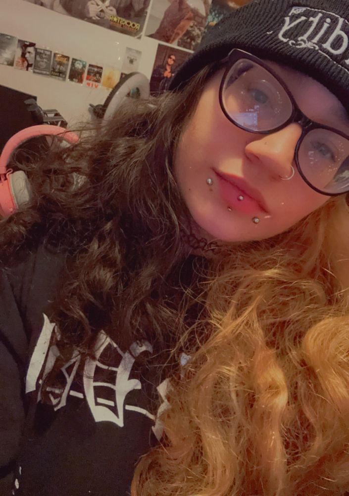 A selfie of Shan. They are wearing a black beanie, black cat eye glasses and a black shirt with white lettering. They have a nose piercing, snakebites and a vertical labret piercing. Their hair is split dyed, brown on one side and blonde on the other.