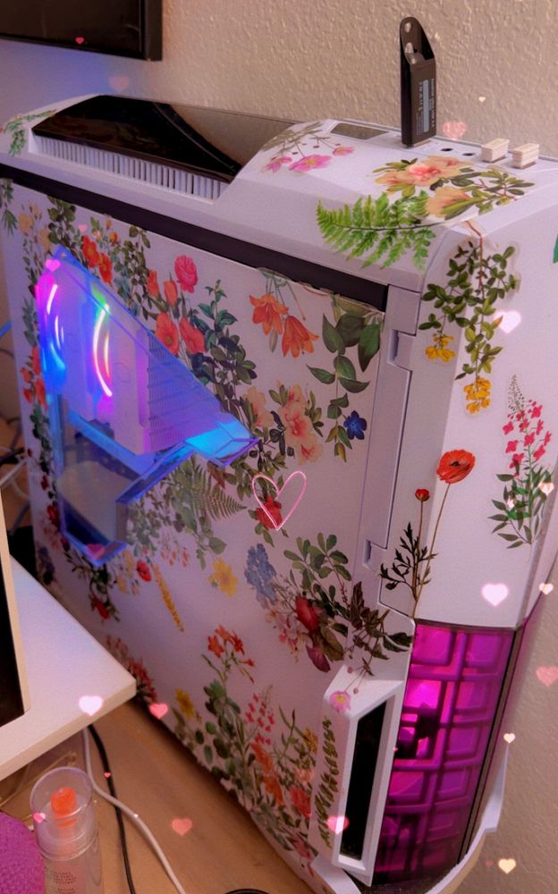 A photo of my computer with a filter over the top with small pink hearts raining down. The computer itself has a white case with a triangle cutout to see inside. There are many foliage and flower stickers decorating the outside of the case. There is a cutout in the front of the case where you can see pink fans running, and through the side window you can see rbg fans. 