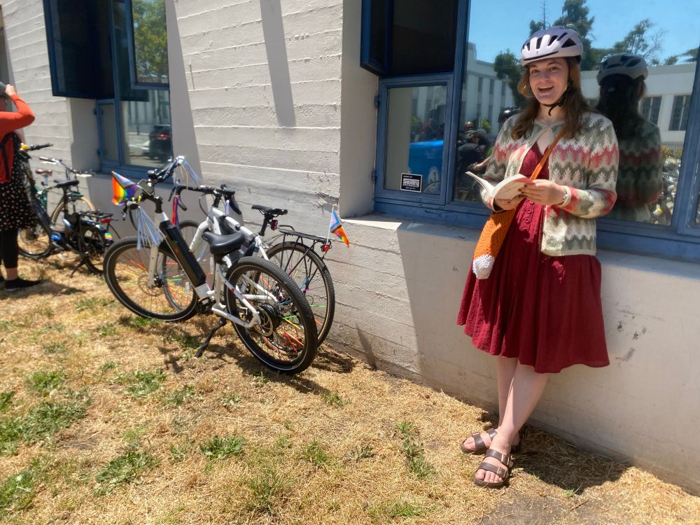 A rider in a dress and helmet leans against the library in a shady spot to read a few pages, near some parked bikes. 