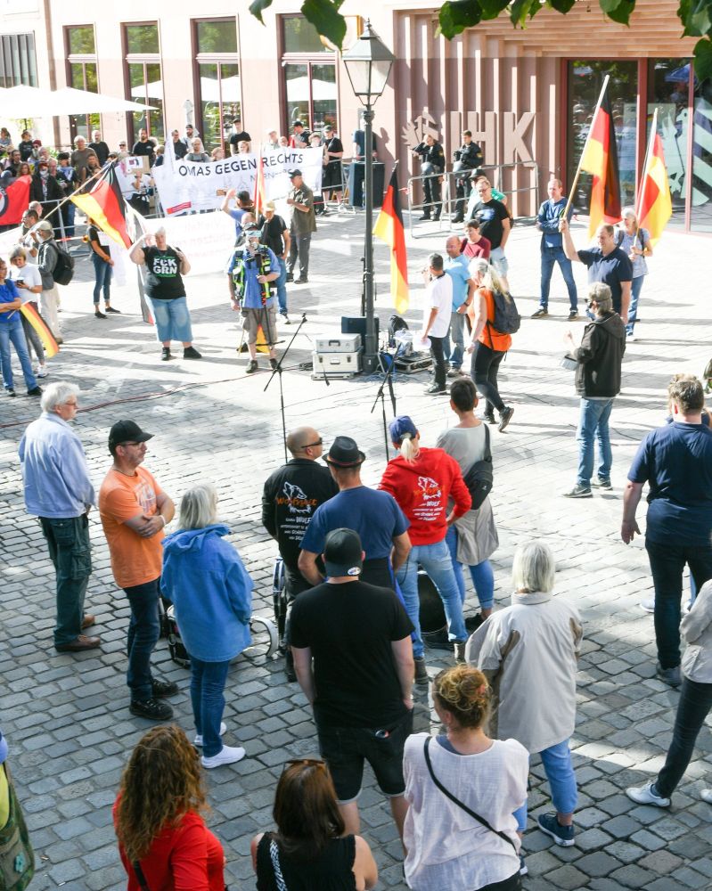 Blick von oben auf die Reste der Versammlung nach Rückkehr von der Demonstration. Im Hintergrund stehen Omas gegen Rechts bei der Gegendemonstration. Zu sehen sind auf der Seite der Rechte noch ein paar Träger mit Deutschland-Flaggen, zwei Personen mit 