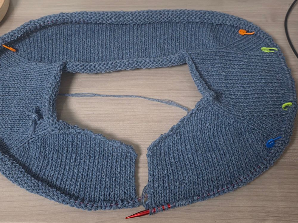 파란색 가디건 편물이 책상 위에 놓여 있으며, 앞부분이 벌어져 있고 직사각 형태로 네크라인이 보인다. 주황색·연두색·파란색 단수표시링이 간격을 두고 꽂혀 있고, 붉은색 대바늘에 실이 연결되어 있다. 이전 사진보다 길이가 늘어나고 형태가 명확해진 상태로, 옷의 어깨에 해당하는 부분을 평면뜨기로 뜨고 있음을 알 수 있다.