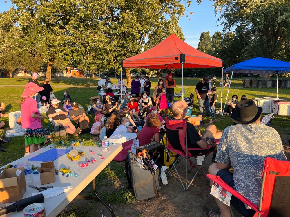 Photo shows people sitting & enjoying performances (magic show by Travis Bernhardt, live music by Geoff Berner). All are wearing respirator masks. It’s a sunny day in a park. There’s a table in the foreground, with colourful craft materials to decorate masks with. The table is abandoned now as the children are enthralled by the magic show.