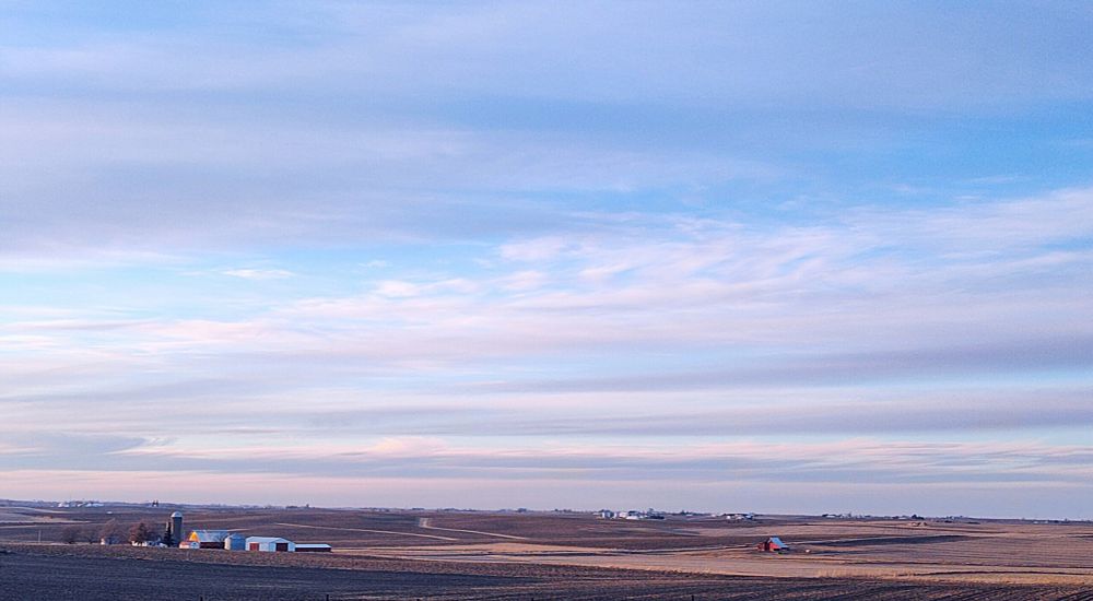 A picture of a bare, brown Midwest cornfield, a farm with a silo and white out buildings in the distance to the left, and a red barn in the distance to the left. The brown horizon meets a bright blue sky peeking through white and shades of grey cloud cover.