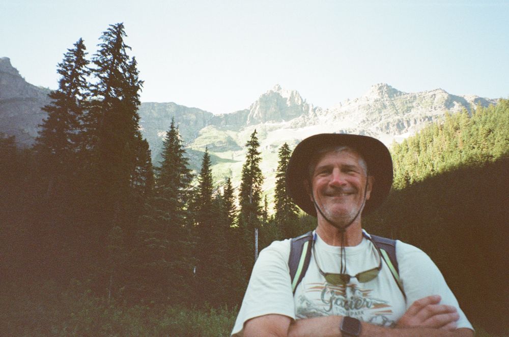 a man (my dad!) in a brimmed hat and glacier tee shirt stands with his arms crossed in front of towering pine trees 