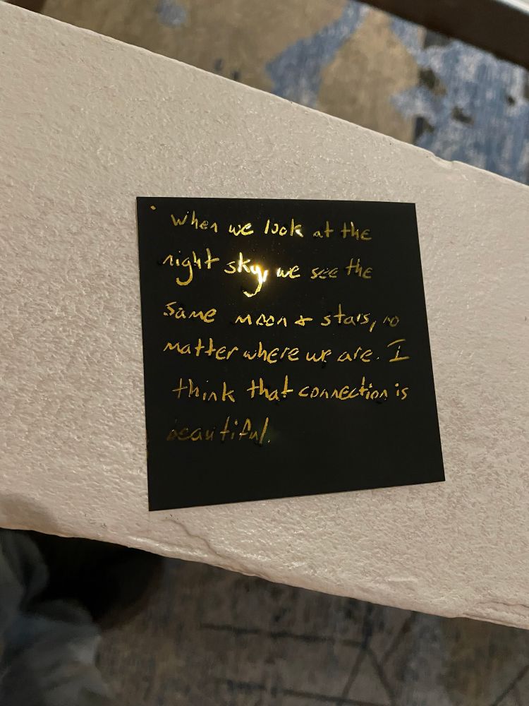 Photo of a small, square, black card placed on a ledge, with some of the black coating scratched away in the shape of letters to reveal a golden background reading "When we look at the night sky, we see the same moon & stars, no matter where we are.  I think that connection is beautiful."