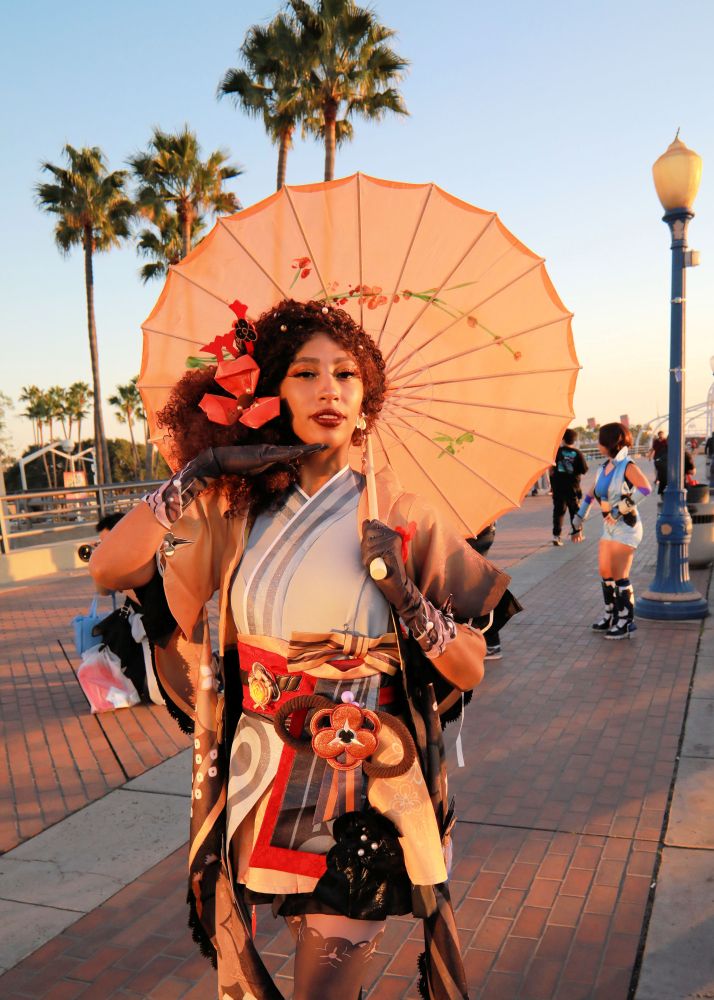 Young woman in a blue wrap dress holding a tan parasol over her left shoulder.  There are orange colored flowers and bow in her long brown hair.  She has a brown shoulder cover and gloves that are lacey.