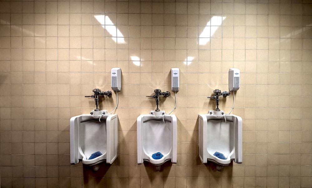 Three porcelain urinals against a yellow tile wall with three scent dispensers above and right of each. Blue splash guards contrast against the white porcelain.  Chrome plumbing shines in the fluorescent lighting.  Four fluorescent lights are reflected against the yellow ceramic 1x1 tiles. 
