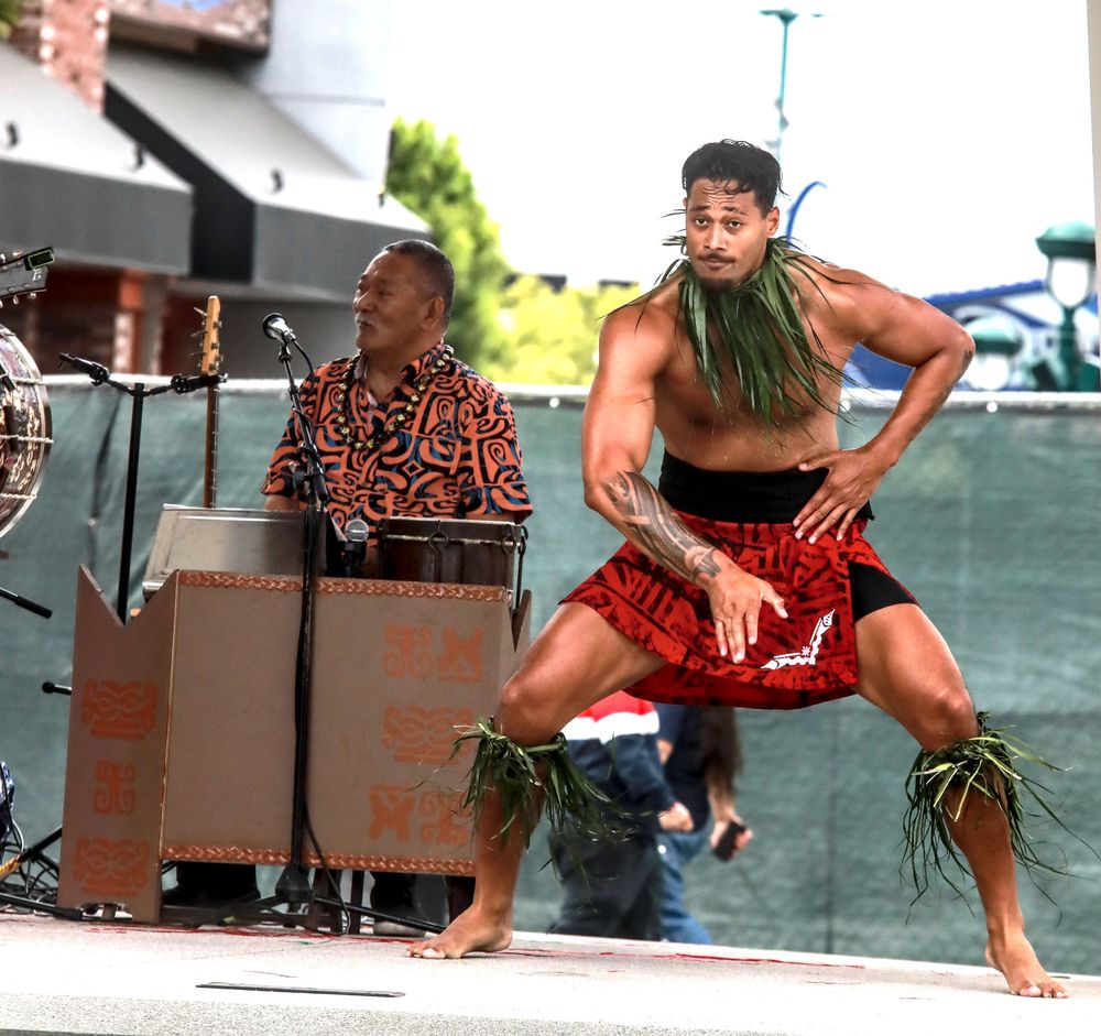 A polynesian dancer from Tupua Productions wearing traditional garb.  The young man is very fit and oiled up, accentuating the muscular physique.  Grass is tied around his neck and just below each knee.  His waist in covered in a red skirt with black trunks underneath.  