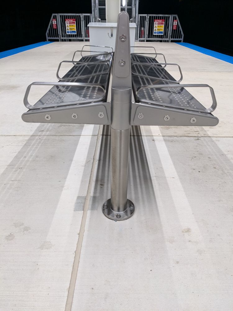 A two-sided steel bench on a concrete train platform. A low back divides it lengthwise, four slim arms divide it into three sections, and the seats slant forward about 15 degrees.