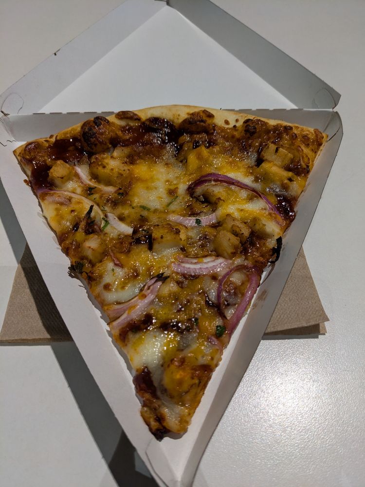 A large triangular New York style slice of BBQ chicken pizza in a triangular slice box, with a brown paper napkin under it, on a white table.