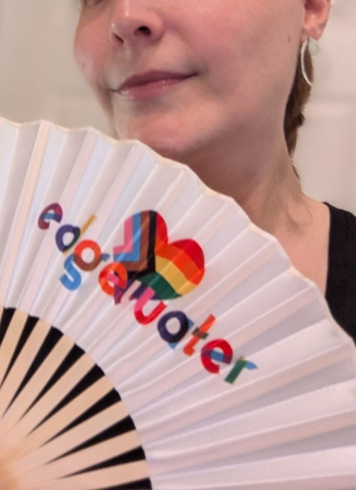A white fan with tan bamboo spokes held just below the lower half of a smiling person's face. The face of the fan is decorated with a heart-shaped Pride flag emblem and the word "Edgewater" in chunky multicolor text.