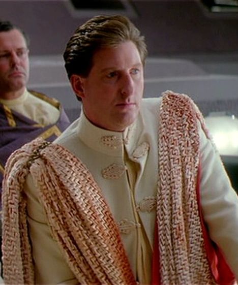Adrian Dunbar sits in a Senate Pod in a regal white jacket with a red and white cloak around him. He has the face of an actor knowing he's going to be a deleted scene trivia answer