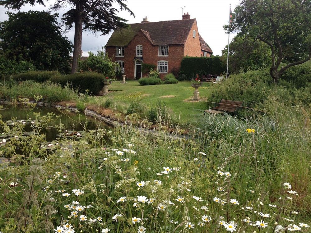 A large, red brick house set within the grounds of a beautiful garden, featuring wildflowers and a wildlife pond