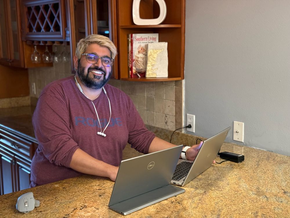 Subject is in a picture using a marble kitchen counter as a work area with multiple monitors and a laptop. Kitchen decor is set to aughts Tuscan farm house style.