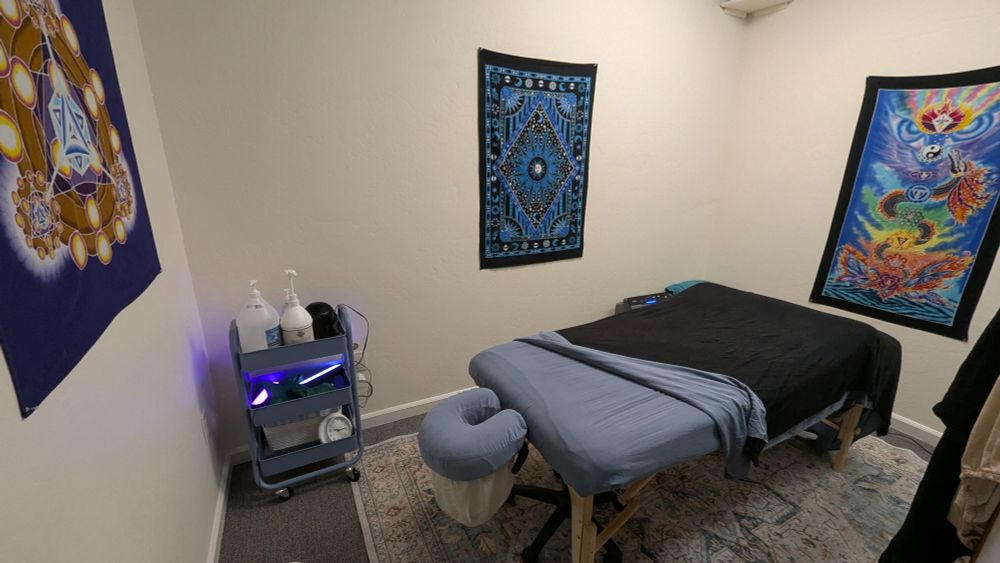 Inside of massage room with a fully set massage table, with sheets and blanket.  Batik wall hangings and a cart with massage oil and supplies. 