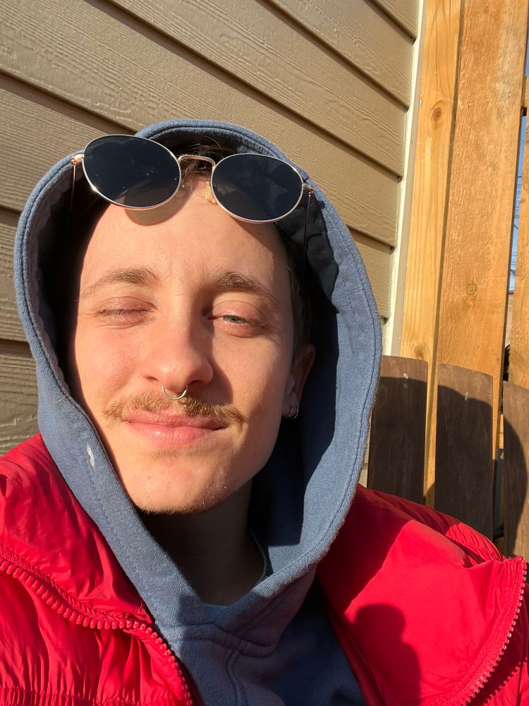 The face of a white person in the sun. They have a mustache. Wearing a blue hoodie and red jacket. 