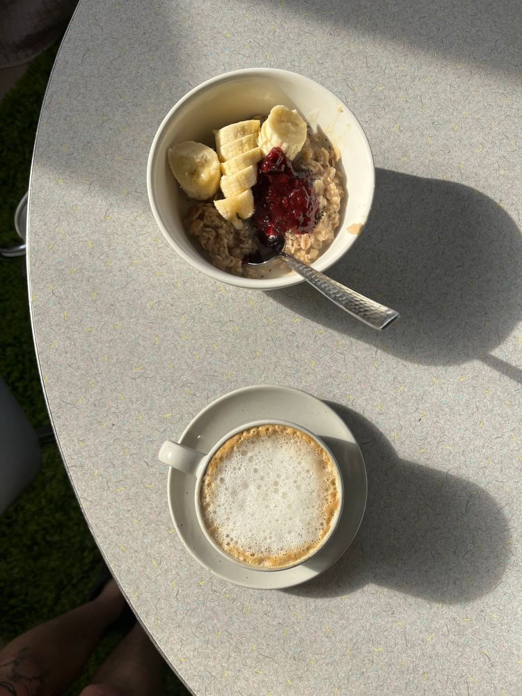 a bowl of oatmeal and a cup of coffee in the sunlight. the oatmeal has bananas and jam. 