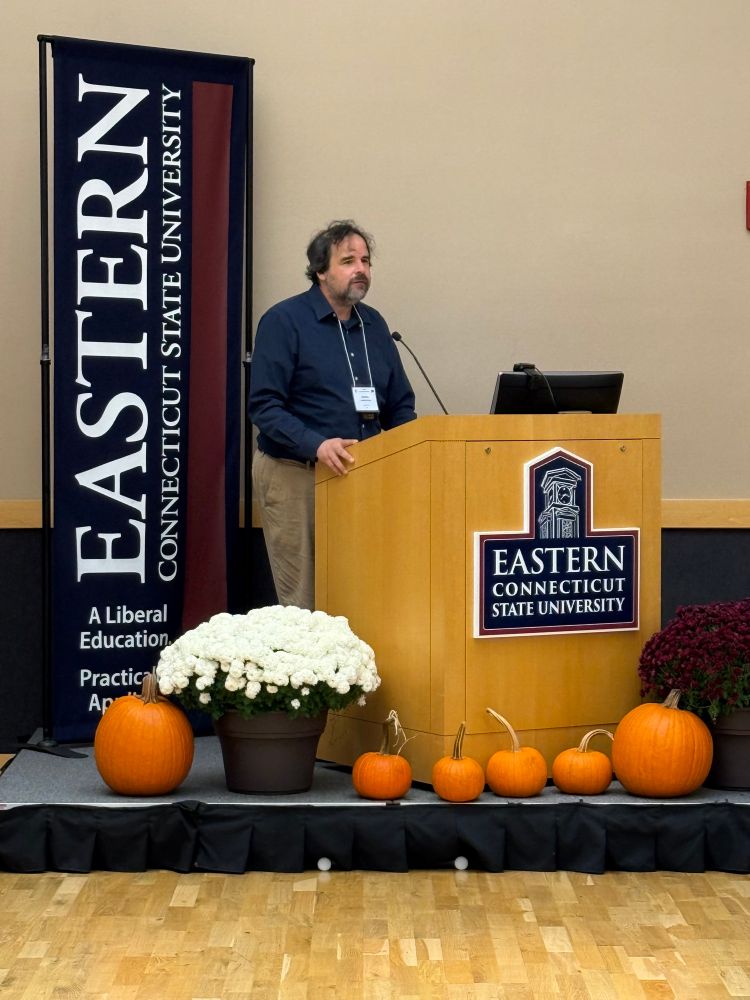 Author Matthew Hongoltz-Hetling speaks at a podium flanked by ECSU signs, pumpkins, and mums. 