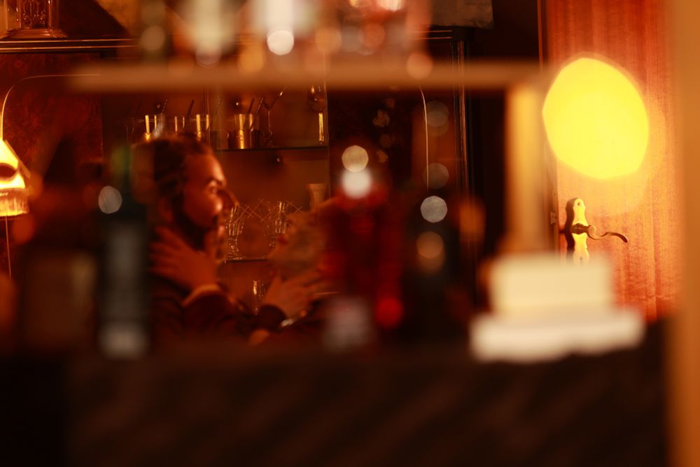 In this photo we can see the foreground blurry as intended. The background is in focus here, with a glass cabinet filled with glasses in the back. In the middle ground we can see two people cosplaying Silco and Vander. They are grabbing each other by the throats, Silco is sticking his tongue out dramatically. 