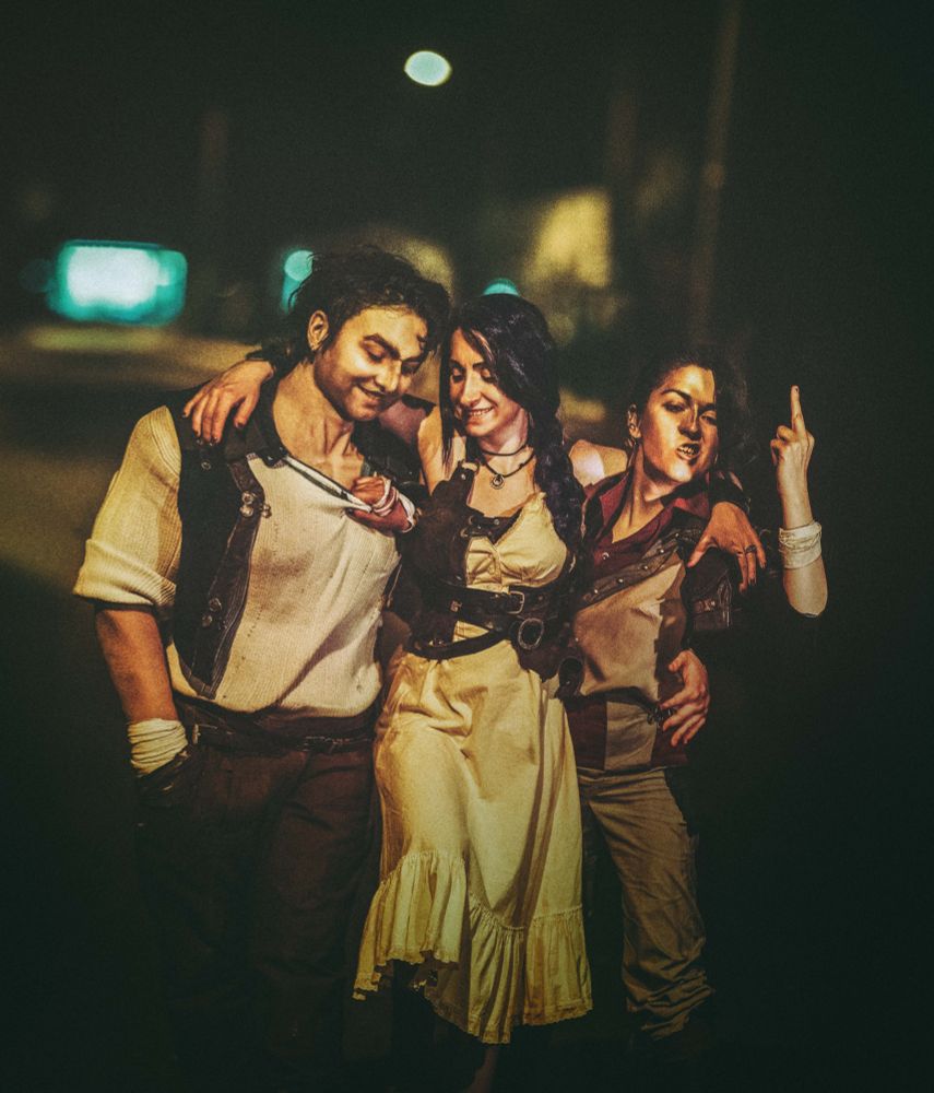 Cosplay photo of young Vander, Felicia and Silco from Arcane. They are walking down an alley by night, laughing and holding on to each other. Felicia has her arms around both men's shoulders. Silco is giving someone the finger, and grabbing Vander's shirt with the other hand. Vander's arm is reaching around both, to grab Silco by his waist. The photo is recreating a piece of fanart by jujudisegnacose