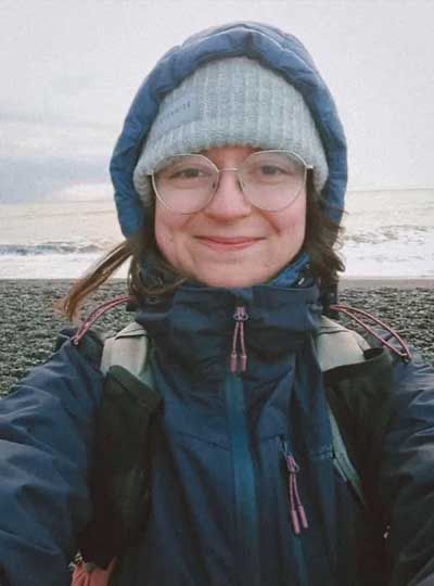 Becky Burke wearing glasses and an anorak takes a selfie in front of the sea