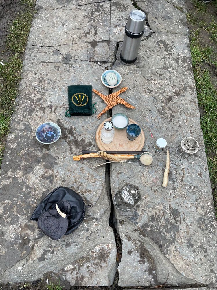 Imbolc altar on a cracked stone slab in the ground showing various objects including a Brigid’s Cross, lantern, wand and Awen symbol.