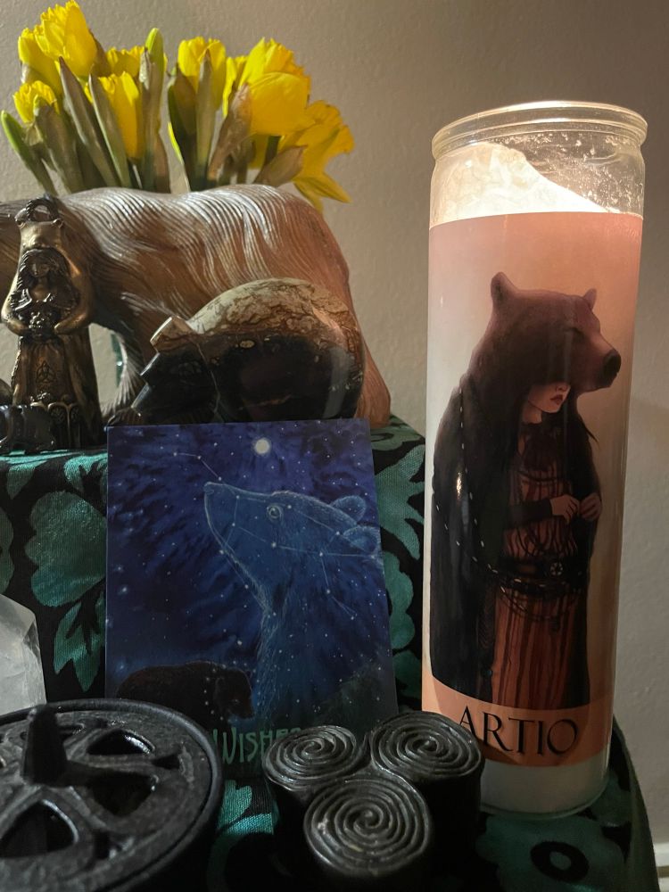 An altar with an offering candle depicting a women wearing a bear mantle and the name Artio (the Celtic bear goddess) printed at its base.
The altar objects include various carved bears in wood and stone, pictures of bears and offerings of water, incense and fresh spring daffodils.
