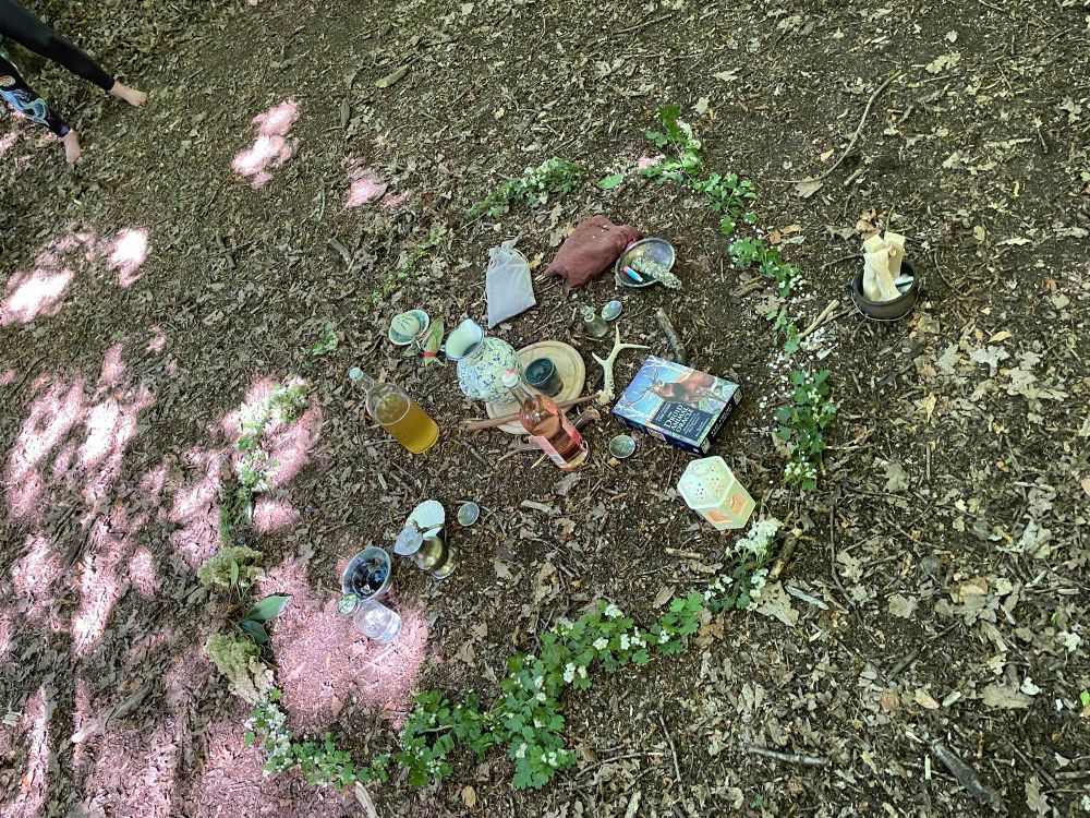 Druid altar on the woodland floor wreathed by hawthorn blossoms. Within, a lit lantern; bowl of water; decorated stone and smouldering herb bundle mark the quarters 