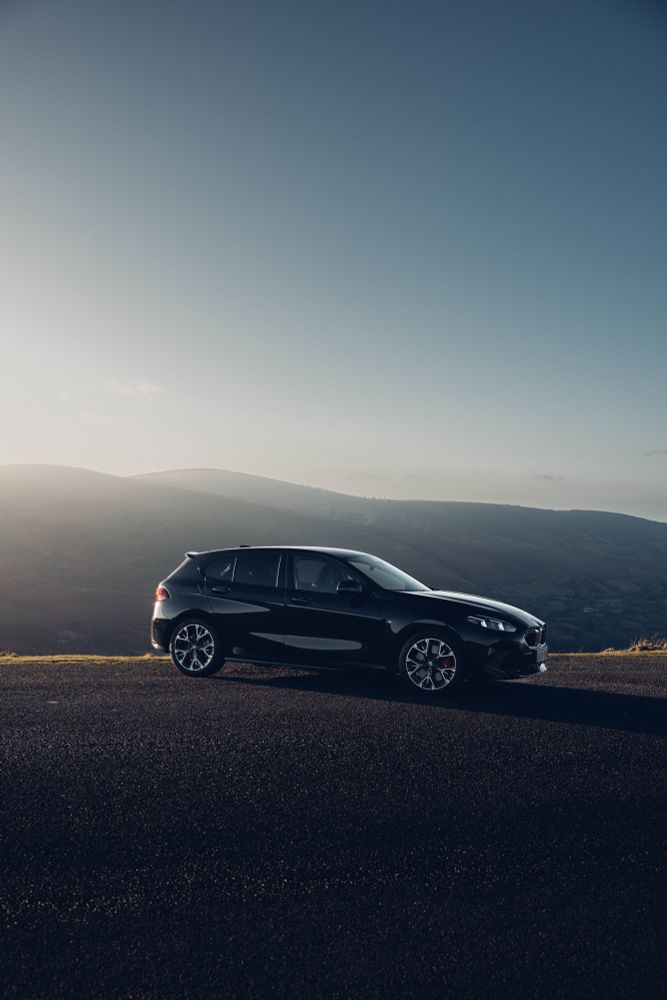 The new BMW 1 Series, photographed from a side seven-eights angle. Low sunshine coming from the left hand side of the photo, casts a long shadow on the ground. Mountains can be been in the distance. 