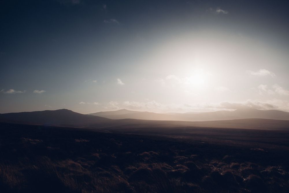 The open plains of the Wicklow Mountains, photographed into strong sunlight. The land is mostly silhouetted, with grass and vegetation just visible in the foreground. 