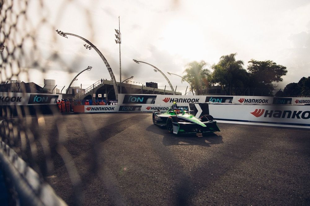 The green Envision Racing Formula E car can be seen cornering through the meshing of the catch fence. 