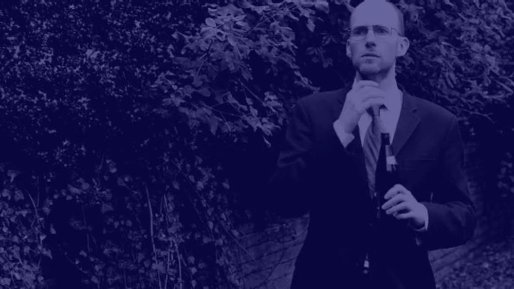 A tall, bald, bespectacled 40-something in a business suit, standing in a street near some bushes, holding a bottle of beer and looking worried