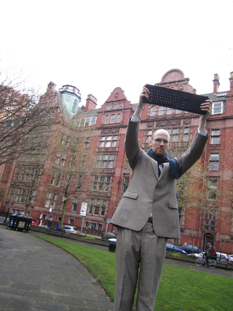 A thin, bald white man in a business suit stands outside the University of Manchester holding a 1980s computer keyboard aloft