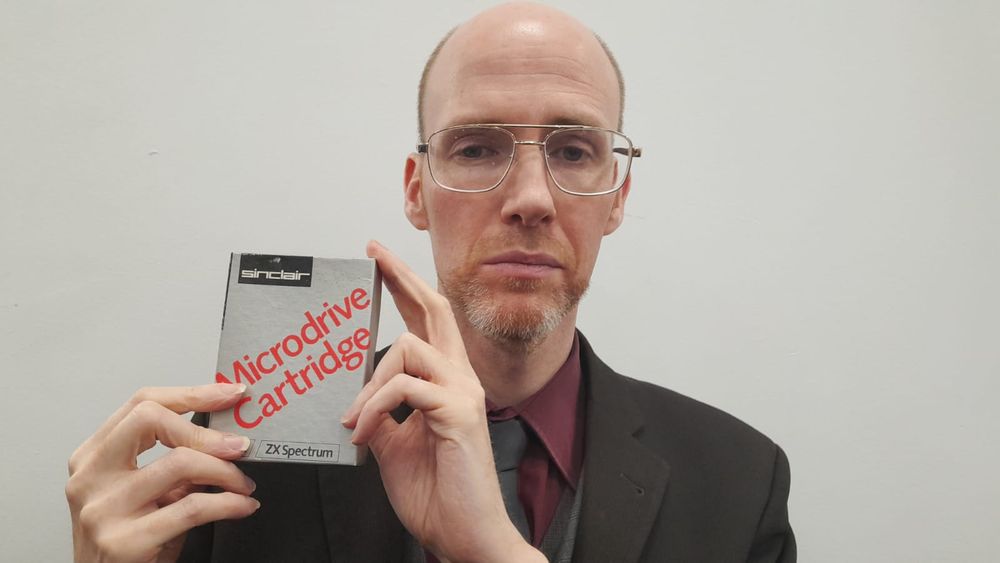 A bald, white, bearded man wearing glasses and a business suit, holding a 1980s Sinclair Microdrive computer cartridge.