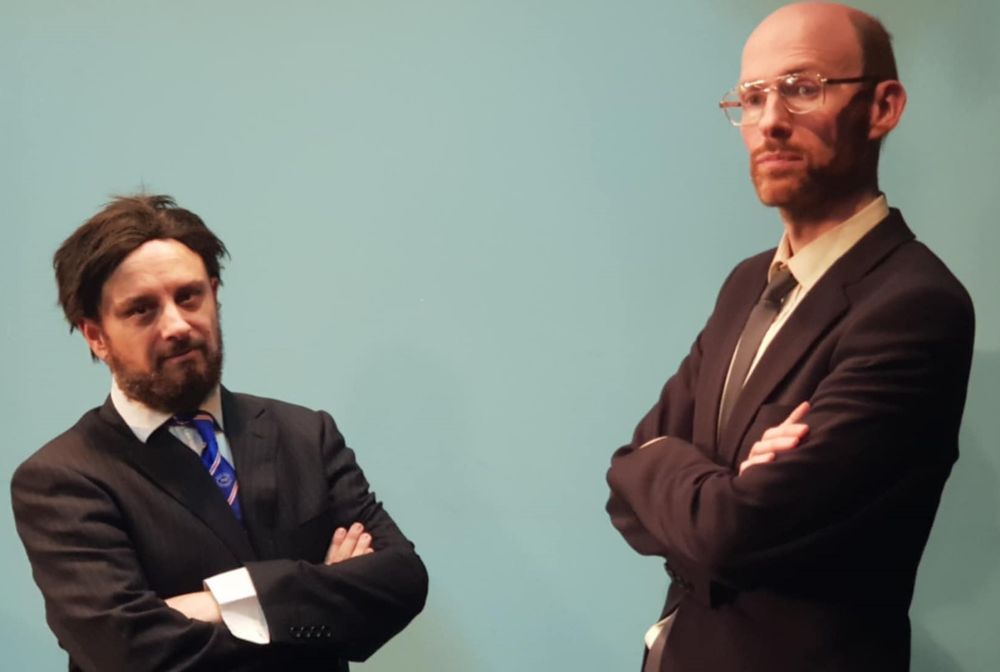 Two serious-looking men in business suits, with their arms folded, looking sternly towards the camera.