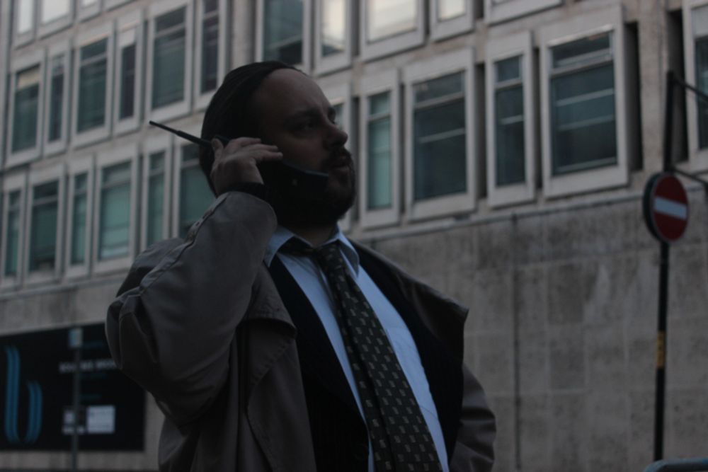 A middle-aged man in a raincoat and business suit standing in a city street talking into a 1980s-style mobile phone.