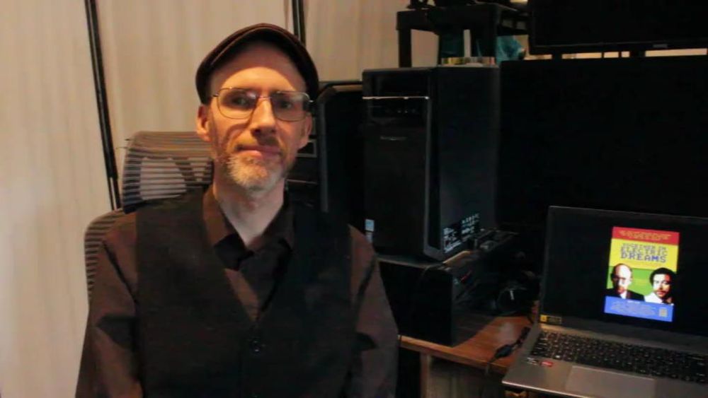 A smiling man in his 40s wearing a flat cap, sitting at a computer desk.